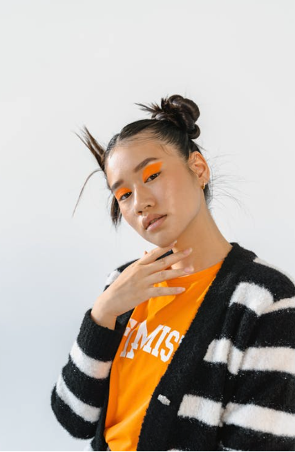 Person with two top buns and bold orange eye makeup wearing a striped sweater and orange shirt, posing with one hand near their chin against a plain background.