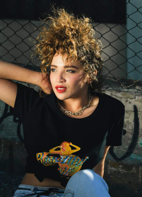 A person with curly hair leans against a graffiti-covered wall, wearing a black T-shirt with a colorful graphic and a necklace.