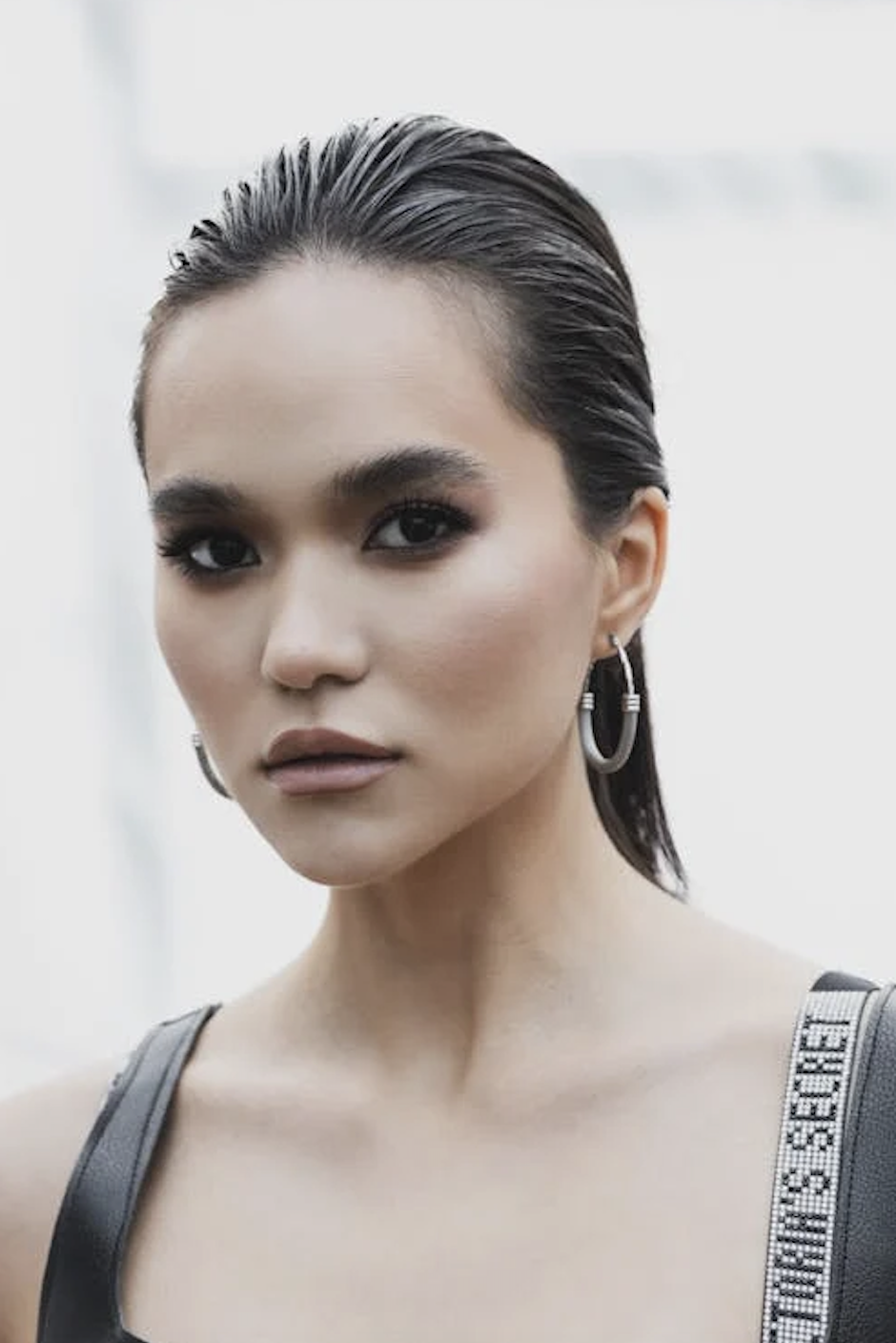 A person with slicked-back hair, hoop earrings, and makeup gazes directly at the camera. They are wearing a strappy garment with decorative elements.