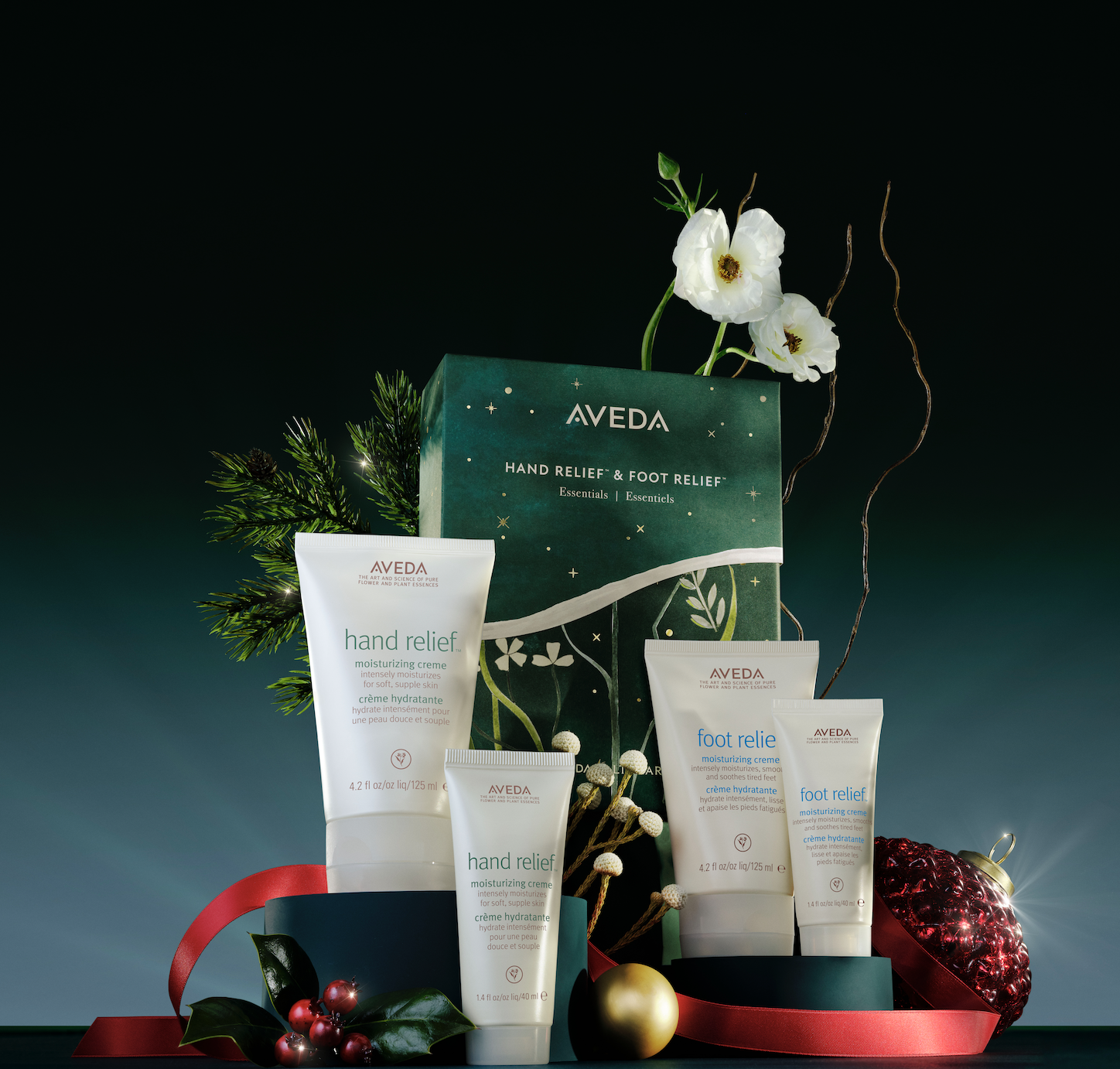 Aveda gift set with hand and foot relief products, decorated with a red ribbon, festive ornaments, and white flowers against a dark background.